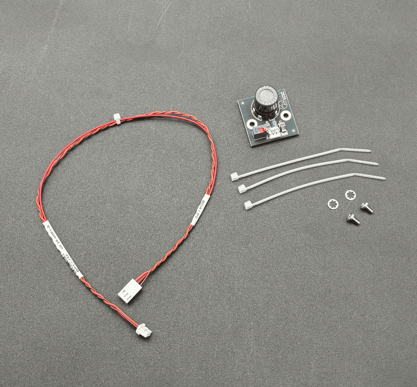 A small electronic control module kit on a dark gray textured background, including a red and white wire harness with a connector, a small circuit board with a rotary knob, three white zip ties, two screws, and two washers.