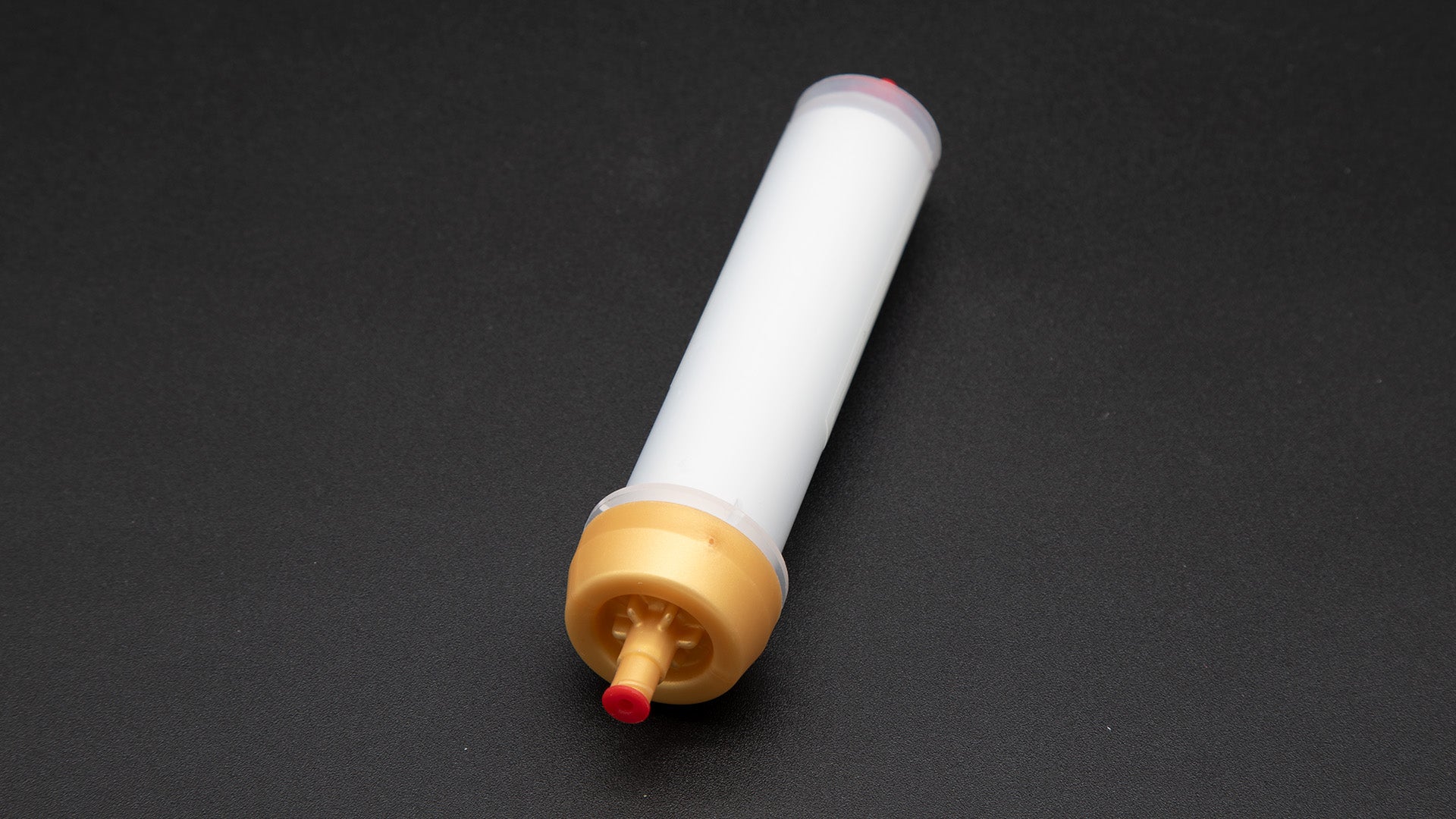 A small white tube with gold and red caps displayed on a black background, creating a striking visual contrast between the two elements.