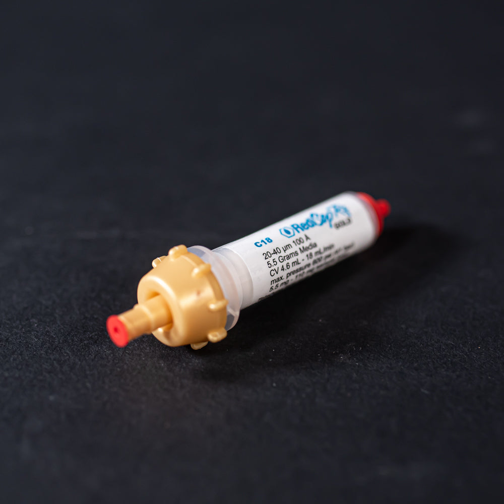 A small white tube with gold and red caps displayed on a black background, creating a striking visual contrast between the two elements.