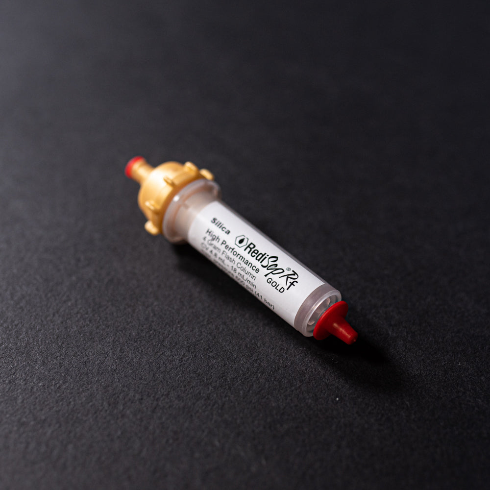 A small white tube with gold and red caps displayed on a black background, creating a striking visual contrast between the two elements.