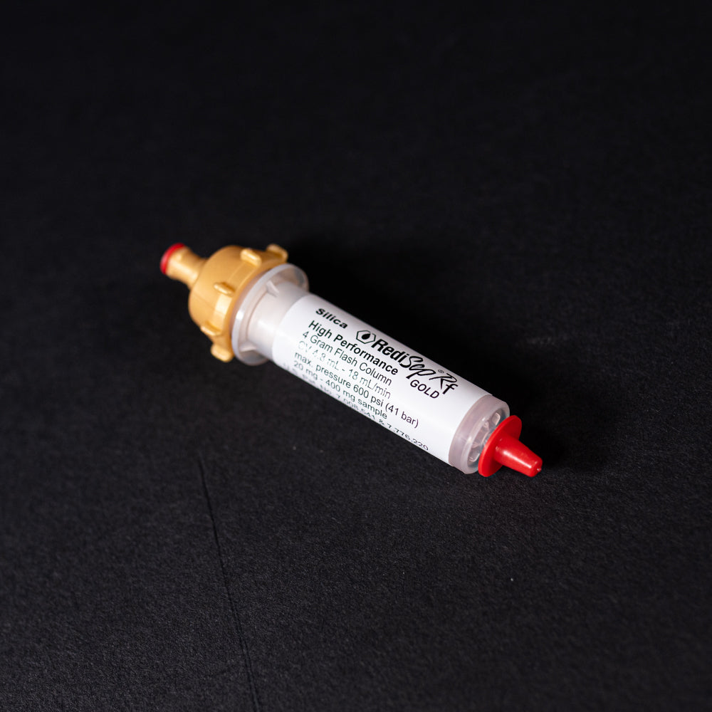 A small white tube with gold and red caps displayed on a black background, creating a striking visual contrast between the two elements.