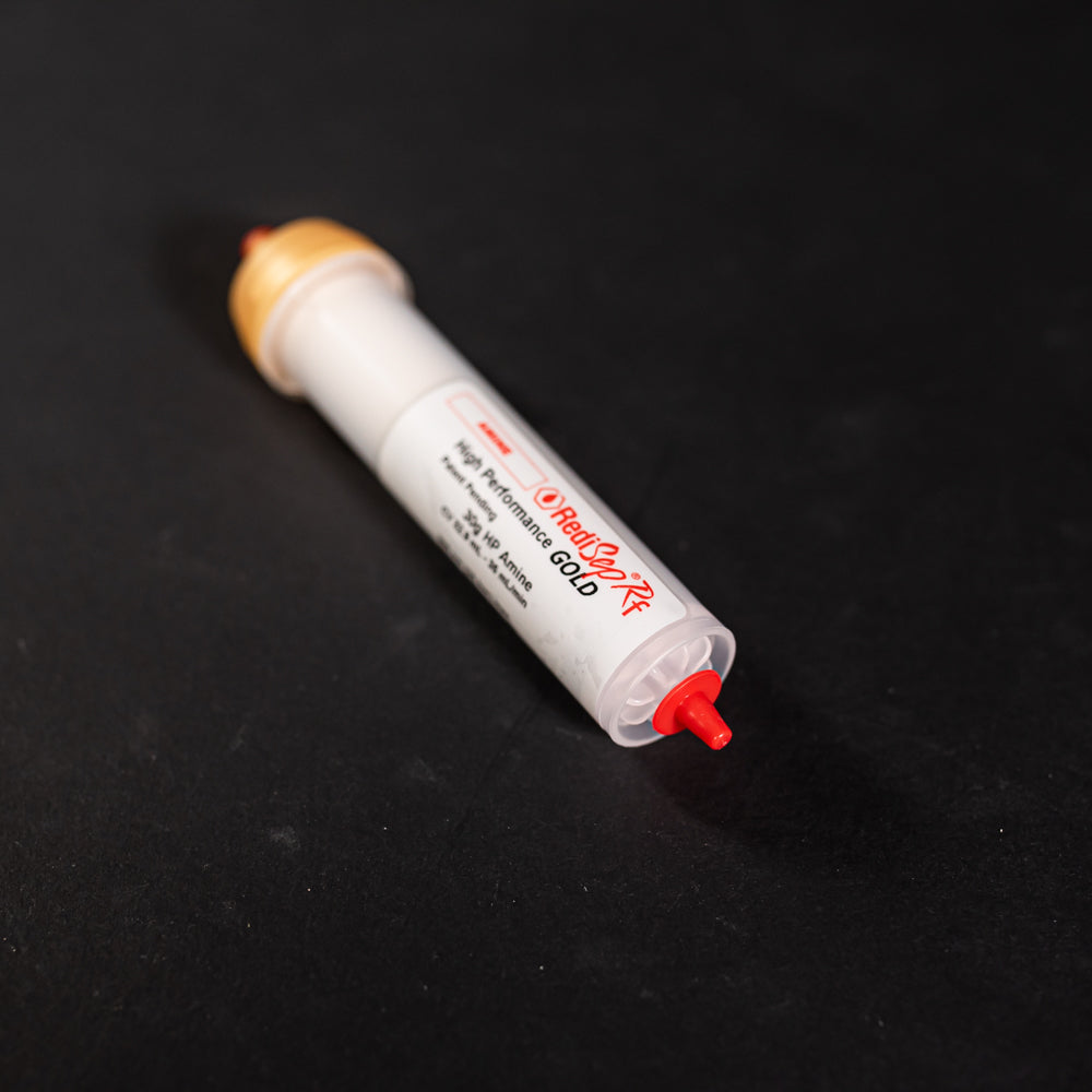 A small white tube with gold and red caps displayed on a black background, creating a striking visual contrast between the two elements.