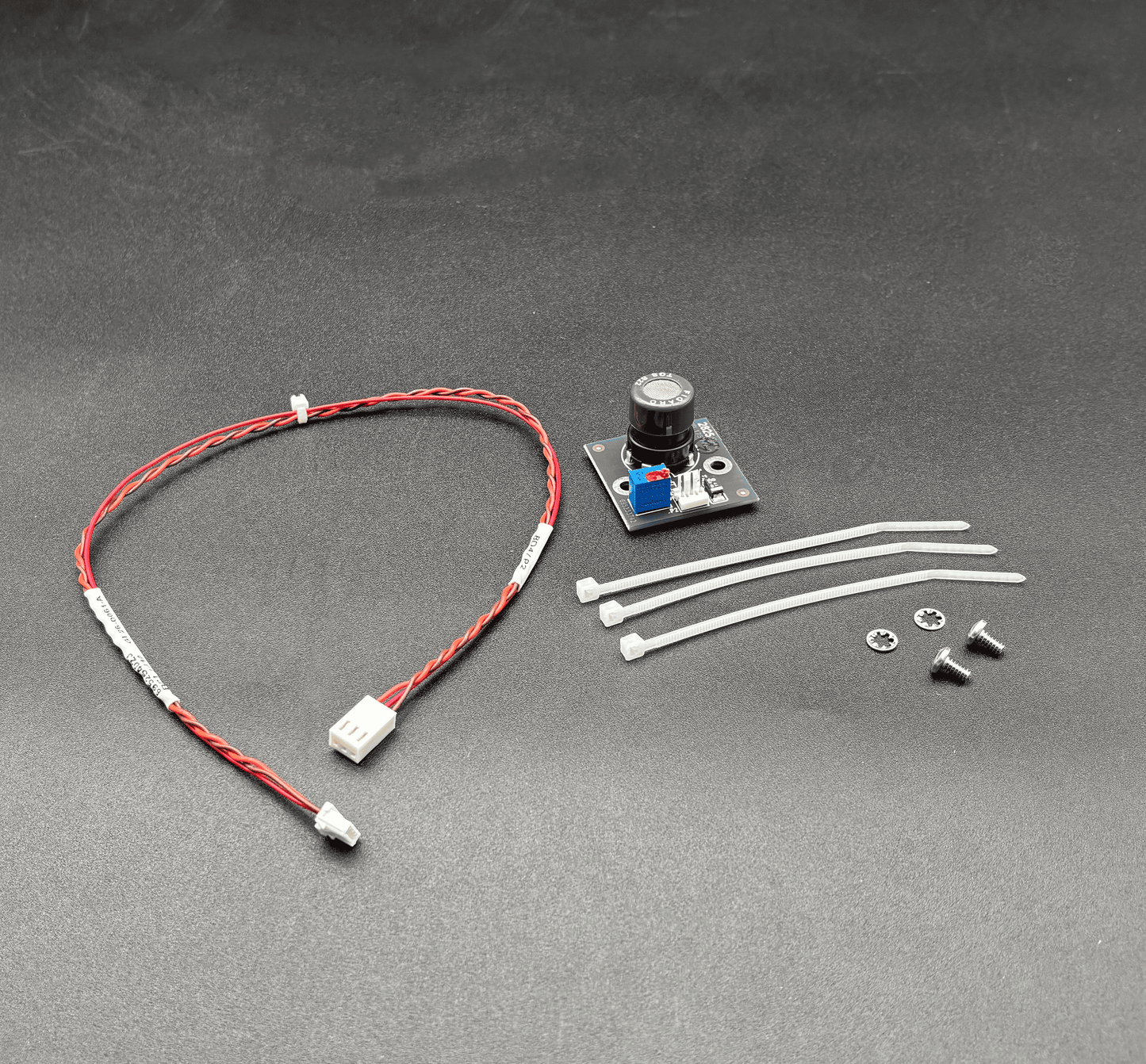 A small electronic component kit on a dark gray textured background, including a red and white wire with white connectors, a small circuit board with a black cylindrical component, four white zip ties, and two small screws.