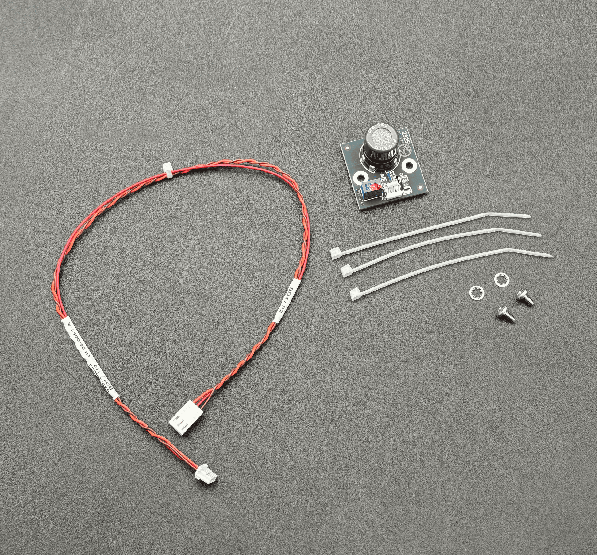 A small electronic control module kit on a dark gray textured background, including a red and white wire harness with a connector, a small circuit board with a rotary knob, three white zip ties, two screws, and two washers.